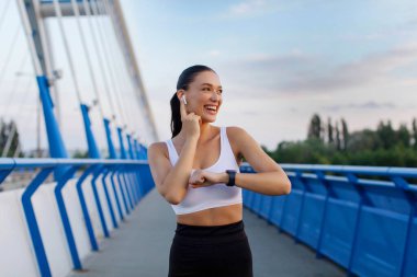 Spor kıyafetleri içinde formda bir bayan antrenmana hazırlanıyor, bayan koşucu dışarıda antrenman yaparken müzik dinliyor, şehir köprüsünde duruyor.