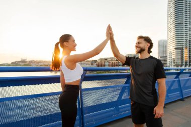 Çift spor başarısı ve takım desteği için beşlik çakıyor, başarıyı kutluyor, egzersiz golleri kazanıyor, gün batımında şehir köprüsünde poz veriyor.