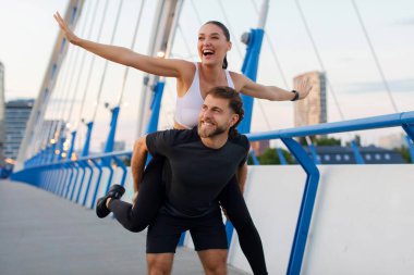 Romantik spor çifti başarılı bir spor antrenmanından sonra eğleniyor, erkek sırtına biniyor ve birlikte gülüyor, şehir köprüsünde duruyor.