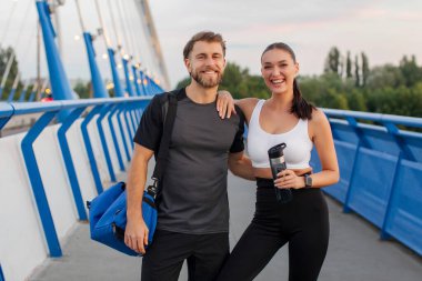 Açık hava eğitiminden sonra birlikte poz veren zinde bir çift, kameraya sarılıp gülümseyen bir adam, spor çantası ve su şişesi tutan bir kadın.