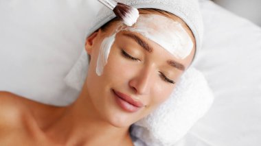 Güzellik uzmanları güzellik salonundaki sulu cilt için kadınların yüzüne vitamin kremi maskesi takıyor. Eldivenli kozmetik uzmanı maske takmak için kozmetik fırçası tutuyor.