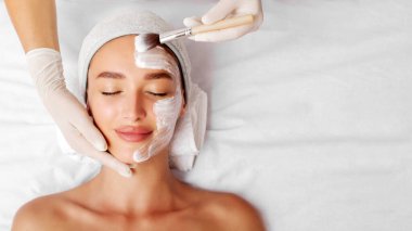Güzeller güzeli genç bir kadın spa salonunda besleyici bir yüz maskesi alıyor. Kozmetik uzmanı beyaz bir maske takıyor. Yüz güzelliği tedavi konsepti