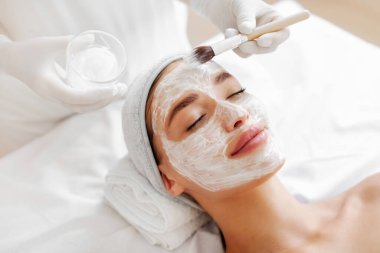 Kaplıcada maske takan, gözleri kapalı yatan ve zevk alan bir kadın. Kozmetik uzmanı kadınların yüzüne fırçayla krem sürüyor.