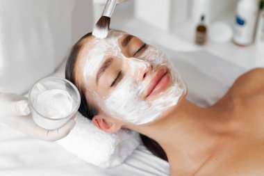 Profesyonel güzellik uzmanı, güzellik salonundaki güzellik prosedürleri sırasında kadınların yüzüne beyaz besleyici maske takıyor, kadın yüz bakımından hoşlanıyor.