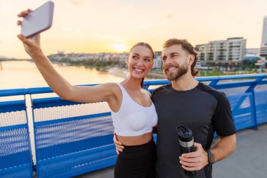 Romantik Avrupalı çift, akşam koşusundan sonra akıllı telefondan özçekim yapıyor ve dışarıda çalışıyor, köprüde duruyorlar.