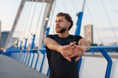 Atletik genç sporcu, kollarını açıyor, kentteki yürüyen köprüde antremana hazırlanıyor, ısınıyor, sokakta koşuyor, boşluğu kopyalıyor.