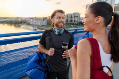 Avrupalı çift dışarıda çalıştıktan sonra dinleniyor, çantalar ve su şişeleriyle ayakta duruyor, sohbet ediyor ve gülümsüyor.