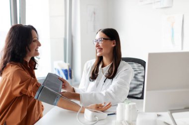 Beyaz önlüklü genç kadın doktor klinikte muayene sırasında dijital tonometre kullanarak orta yaşlı kadın kan basıncını ölçüyor.