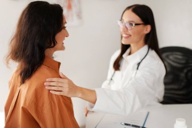Dost canlısı doktor orta yaşlı kadın hastayı destekliyor, elini omzuna koyuyor, klinikte muayene sırasında konsültasyon yapıyor.