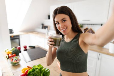 Mutlu bayan sabah antrenmanından sonra smoothie ile poz vererek selfie çekiyor, evde mutfağın içinde duruyor, sağlıkla ilgilenen yeşil detoks içeceğinin keyfini çıkarıyor.