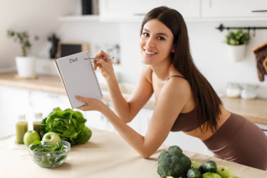 Beslenme planı olan, kameraya gülümseyen, not defterine menü ve yemek tarifleri yazan, mutfakta duran mutlu formda bir bayan. Zayıflama beslenme kavramı