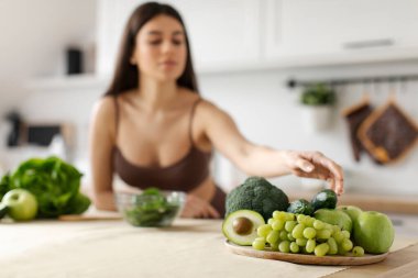 Yeşil sebzeler ve tabakta meyveler üzerine seçici bir odak, modern mutfakta sağlıklı bir akşam yemeği hazırlayan, malzeme alan sağlıklı bir kadın.