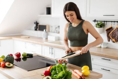Spor giysili formda bir kadın taze sebze salatasına salatalık eklerken mutfağın içinde sağlıklı bir yemek hazırlıyor.