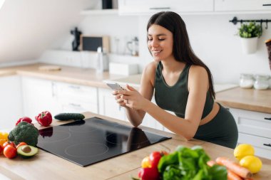 Spor kıyafetli bir kadın cep telefonu kullanıyor, sebze tarifi arıyor öğle yemeği yiyor ve evde sağlıklı bir akşam yemeği hazırlıyor.
