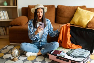 Şok olmuş bir kadın elinde akıllı telefonla turistik kısıtlamalar ya da uçuş iptali hakkında mesaj okuyor, açık bavulun yanında oturuyor.