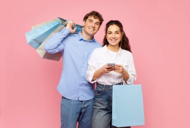 E-ticaret. Renkli alışveriş torbalarıyla mutlu Avrupalı çift cep telefonu kullanıyor, internetten alışveriş yapıyor ve pembe arka planda kameraya gülümsüyor.