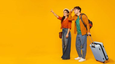 Seyahat teklifi. Seyahat reklamı için fotokopi alanına bakan çift, bavulla ayakta duruyor, yolculuğa hazır, turuncu stüdyo arka planı, panorama