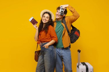 Mutlu turistler tatile hazır, fotoğraf makinesi ve bavullarıyla sarı stüdyo arka planında kameraya gülümsüyorlar.