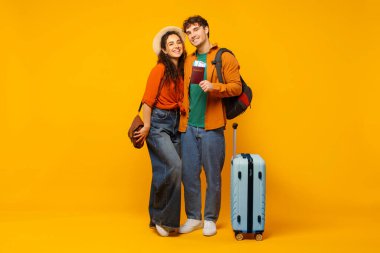 Mutlu eşler tatile hazır, seyahat biletleri ve valizlerle poz veriyorlar, sarı stüdyo arka planında poz veriyorlar, uzun bir çekim.