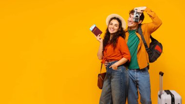 Mutlu turistler tatile hazır, fotoğraf makinesi ve bavullarıyla sarı stüdyo arka planında kameraya gülümsüyorlar.