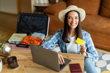 Mutlu bir kadın kredi kartı ve dizüstü bilgisayar kullanıyor, yurt dışına internet üzerinden rezervasyon yaptırıyor ya da evden internet üzerinden otel rezervasyonu yaptırıyor, arka planda açık bavulla oturuyor.