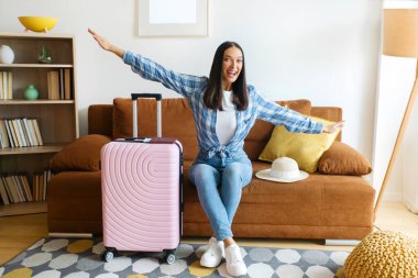 Deniz aşırı seyahat, uçak yolculuğu konsepti. Kanepede oturan neşeli bayan bavulunu hazırlıyor ve uçak jesti yapıyor. Yurtdışına gitmeye hazır.