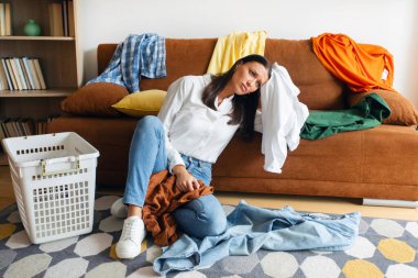 Oturma odasında çamaşırların arasında oturan yorgun kadın yorgun, stresli ve ev işleri programından bıkmış.