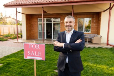 Mutlu orta yaşlı Avrupalı emlakçı yeni evin önünde satılık duruyor, kameraya gülümsüyor, kollarını kavuşturmuş poz veriyor.