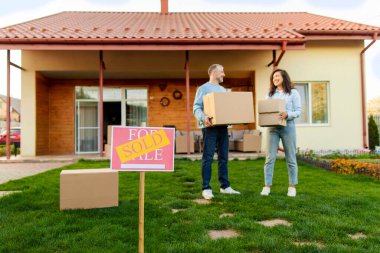 Satılmış tabelalara ve karton kutular taşıyan mutlu eşlere odaklan, arka planda duran evin önünde duran, tam boy