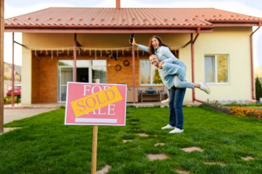 Mutlu eşler emlak satın aldıktan sonra eğleniyorlar, karı koca yeni evlerinin önünde duruyorlar, satılan tabelalara odaklanıyorlar.