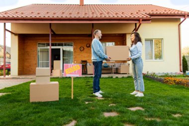 Taşındıkları gün yeni evlerinin önünde karton kutu taşıyan evli bir çift birbirlerine bakıp gülümsüyor. Emlak ve emlak kavramı