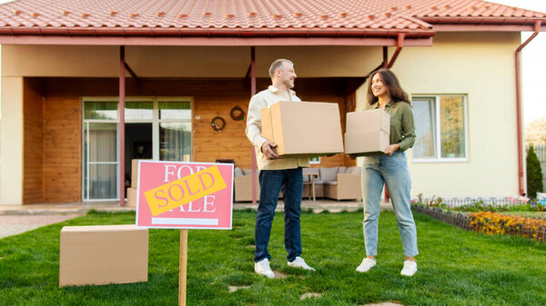 Couple holding boxes standing in front of their home on moving day, focus on sold sign. Real estate and property concept