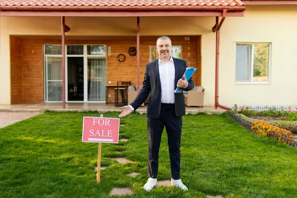 Satılık emlakçı tabelasının yanında duruyor ve güzel bir eve, açık havaya, uzun mesafeli atışlara karşı işaret ediyor.