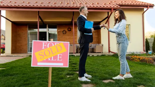 Yeni ev alan genç bir kadın, erkek emlakçıdan anahtarları alan evin önünde ön planda satılan levhalarla duran bir kadın.