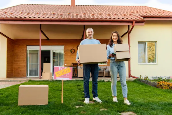 Avrupalı çift taşınma gününde yeni evlerinin önünde kutularla dikiliyorlar. Yakınlarda bir tabela, tam boy, kopyalama alanı var.