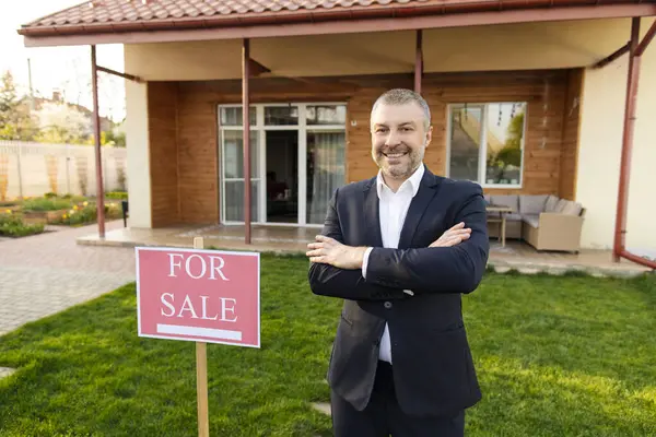Mutlu orta yaşlı Avrupalı emlakçı yeni evin önünde satılık duruyor, kameraya gülümsüyor, kollarını kavuşturmuş poz veriyor.