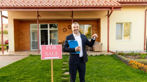 Neşeli erkek emlak yöneticisi anahtarlarını gösteriyor, satılık tabelasının yanında duruyor güzel kır evinin önünde, panorama çekimi