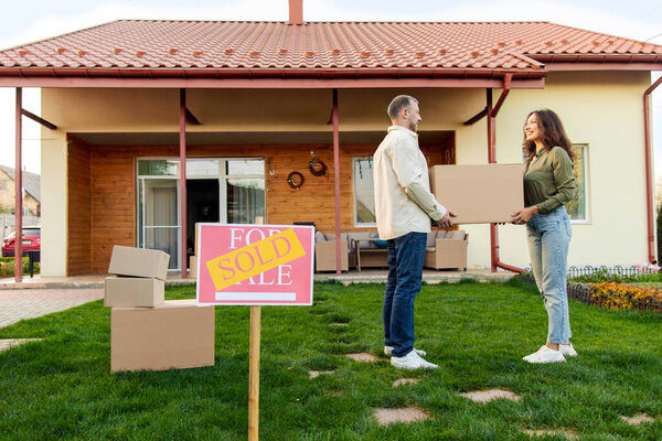 Buying house. Happy spouses with box standing in front of their new home on moving day, SOLD sign on foreground