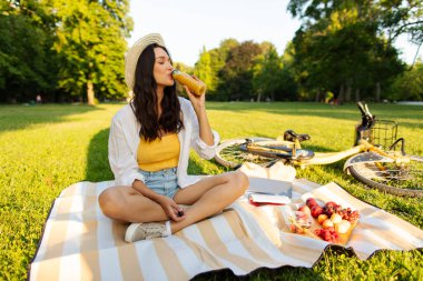Genç bir kadın piknik yapıyor, battaniyeye oturuyor ve portakal suyu içiyor, yaz parkında dinleniyor.