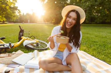 Güneşli bir yaz gününde yerel parkta oturup kameraya gülümseyen ve hazır yemek konteynırından sağlıklı salata yiyen mutlu kadın.