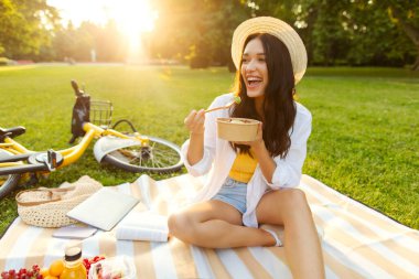 Heyecanlı kadın salata yiyor sağlıklı yemek yiyor, şehir parkında çimlerin üzerinde battaniyeye oturuyor, yaz tatilinde öğle yemeği yiyor.