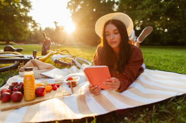 Genç bayan yaz parkında piknik yapıyor ve kağıt kitap okuyor, yakınlarda meyve ve meyve suyu ile battaniyeye uzanıyor.