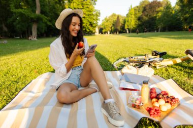 Neşeli bayan ekose üzerinde dinleniyor, cep telefonu kullanıyor, şeftali yiyor, sosyal medyada geziniyor, parkta oturuyor, sağlıklı meyve atıştırmalıkları yiyor.
