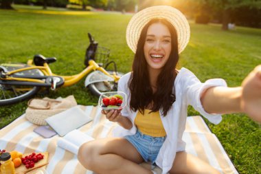 Güzel, mutlu bir bayan selfie çekiyor, şehir parkında piknik yapıyor, yeşil çimenlerin üzerinde battaniyeye oturuyor ve meyve salatası yiyor.
