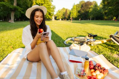 Güzel genç bayan parkta piknik yapıyor, çimlerin üzerinde ekose oturuyor, cep telefonu kullanıyor ve taze meyve yiyor, bedava yer açıyor.