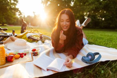 Güneşli bir günde yeşillik parkında piknik yaparken gazete okuyup meyve yiyen rahat genç bir kadın.