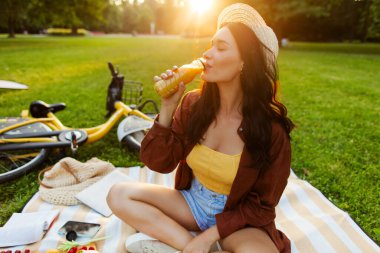 Güzel, rahat genç bir kadın soğuk portakal suyuyla parkta dinleniyor, piknik yapıyor, yanında bisikletle battaniyede oturuyor.