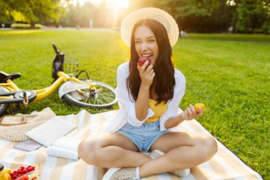 Heyecanlı genç bir kadın yaz güneşli bir günde parkta piknik yaparken meyve yiyor, şeftali veya nektarin tutuyor ve yiyor.