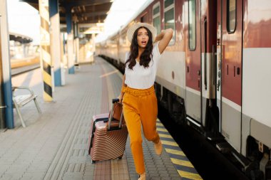 Tren istasyonunda koşan ve giden treni kovalayan bir kadın el sallıyor ve bavuluna binmek için acele ediyor.