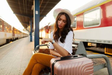 Tren istasyonunda elinde bavulla bankta oturmuş saati seyreden üzgün bir kadın.